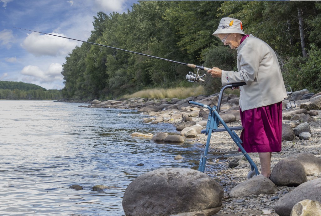 horizontal image of an elderly senior woman with a walker fishing at the edge of the lake with trees and rocks lining the shoreline in the summer time